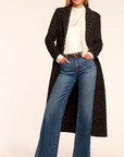 Woman wearing a long black coat, white top, and blue jeans on a plain background