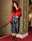 Woman in a red blouse and patterned skirt standing in front of an ornate wall.