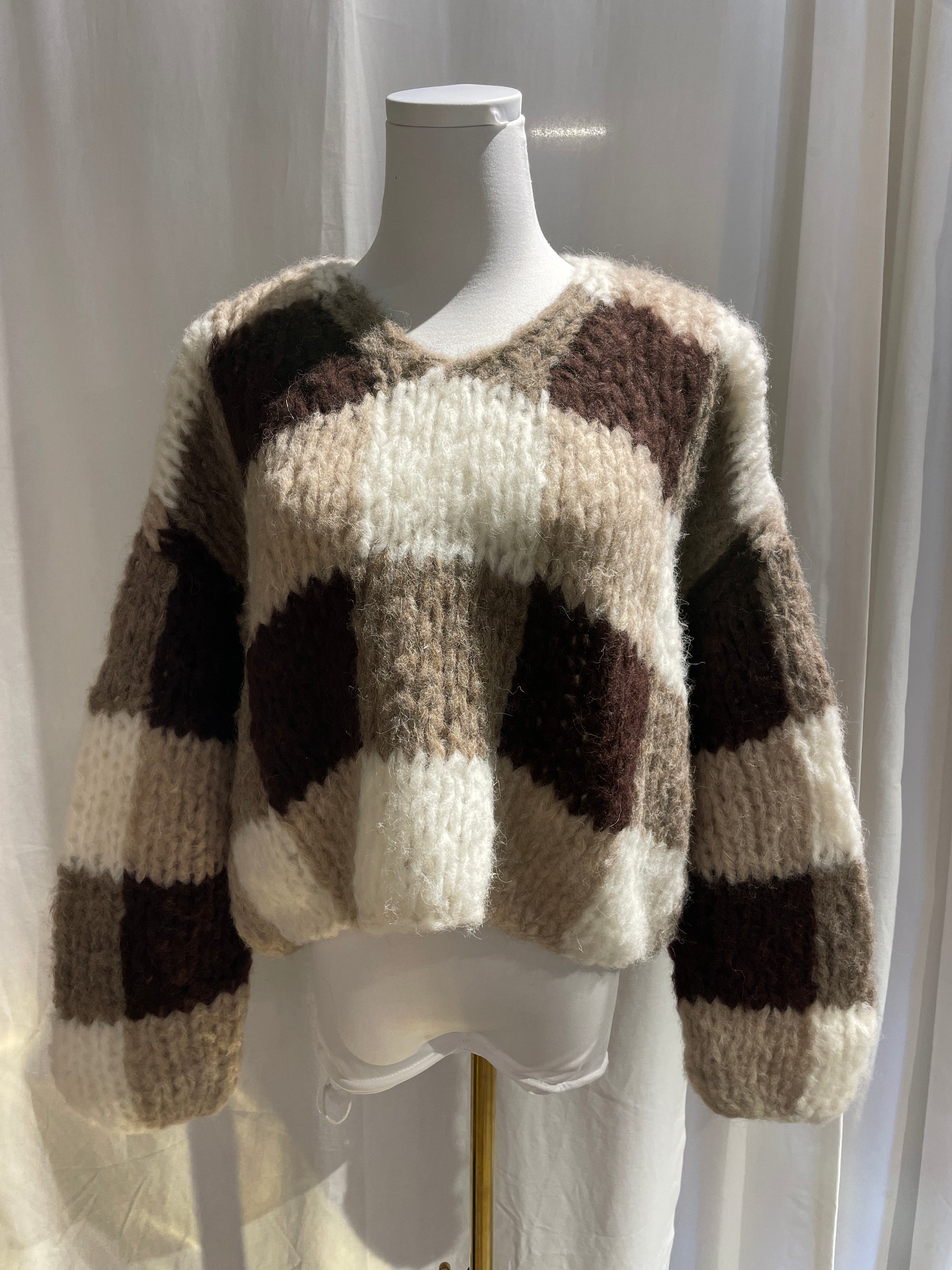 A white mannequin with a white4, beige and brown checkered sweater hanging on it with a white background.