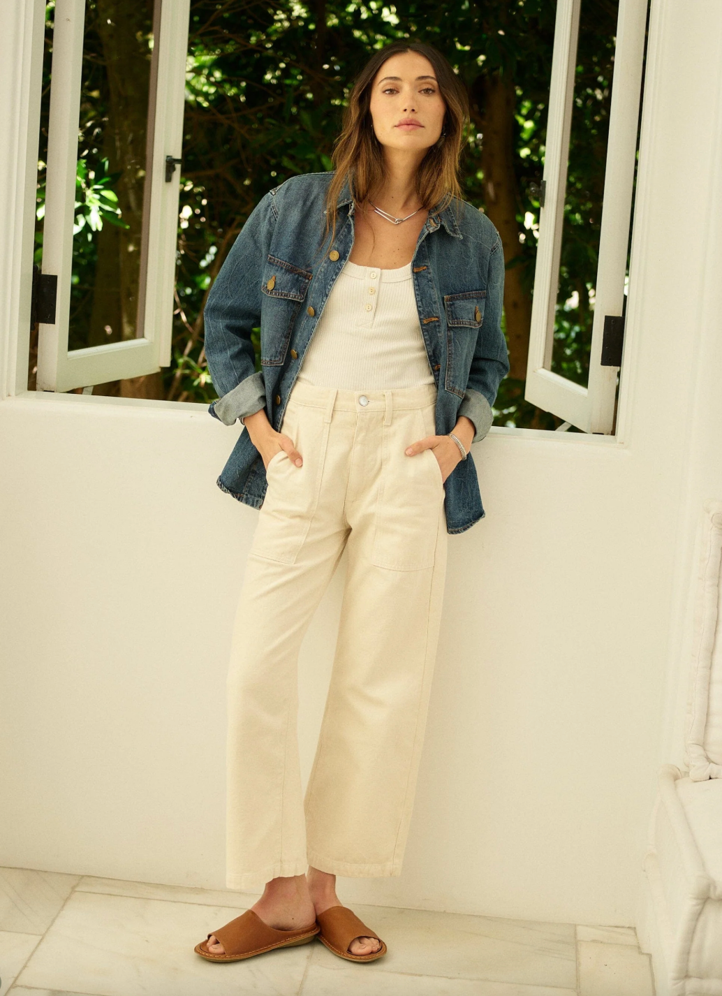 Woman wearing a denim jacket and white pants standing in a bright room with large windows.
