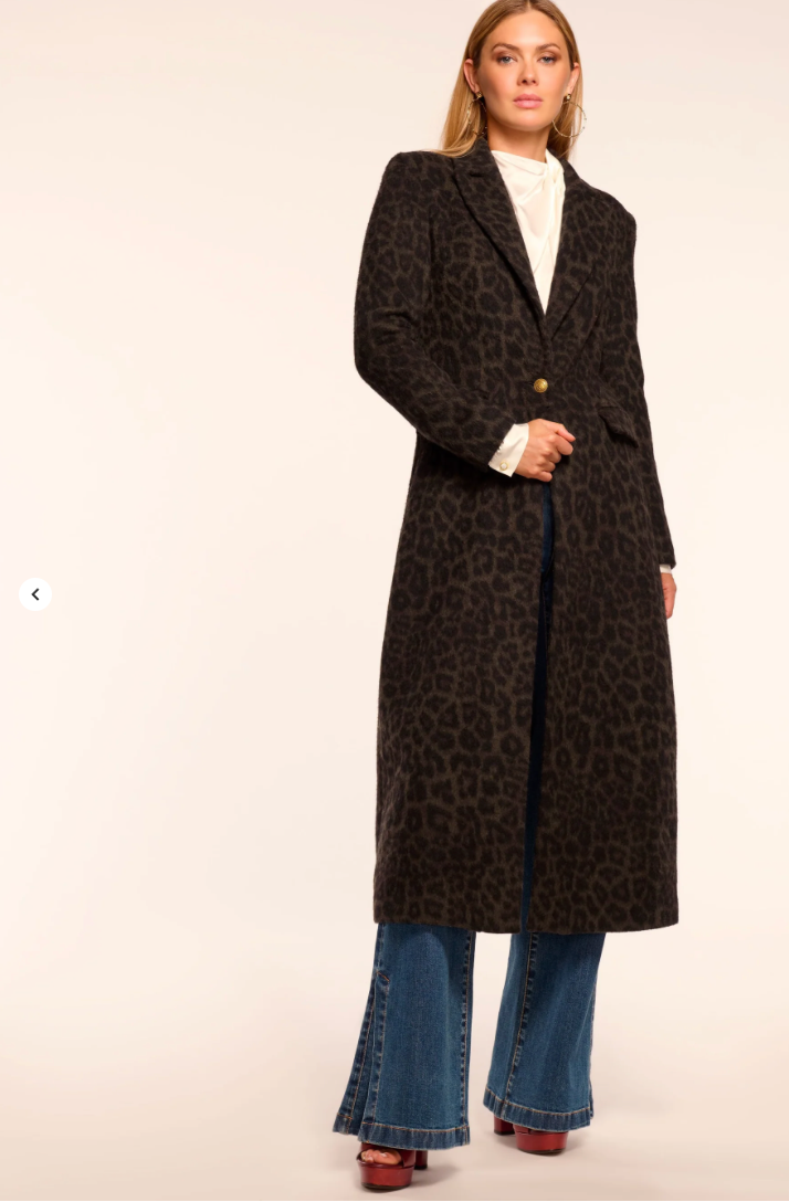 Woman wearing a long dark coat with a pattern, white shirt, and blue jeans on a beige background