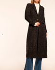 Woman wearing a long dark coat with a pattern, white shirt, and blue jeans on a beige background