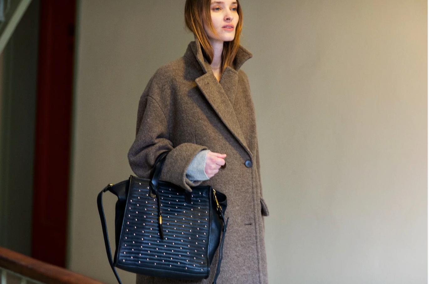 Woman in a brown coat holding a black handbag against a plain background