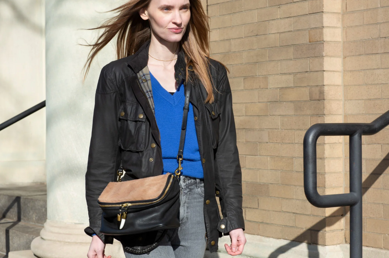 Woman wearing a black jacket and blue shirt with a brown bag, standing against a brick wall.