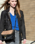 Woman wearing a black jacket and blue shirt with a brown bag, standing against a brick wall.