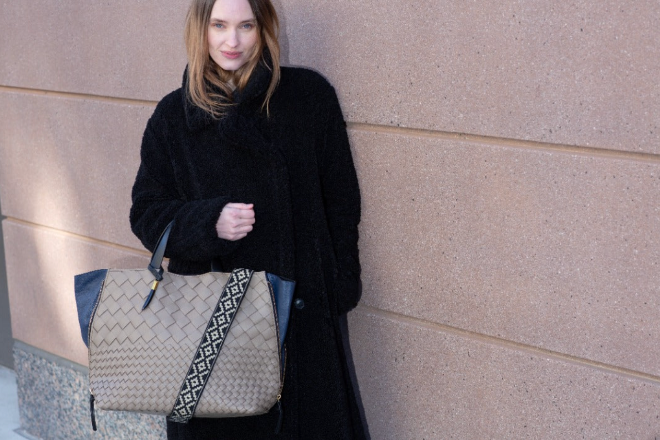 Woman holding a beige handbag with a patterned strap against a beige wall.
