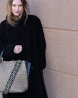 Woman holding a beige handbag with a patterned strap against a beige wall.