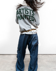 Person wearing a hoodie with 'Artists Wanted' text on the back, standing against a plain background.