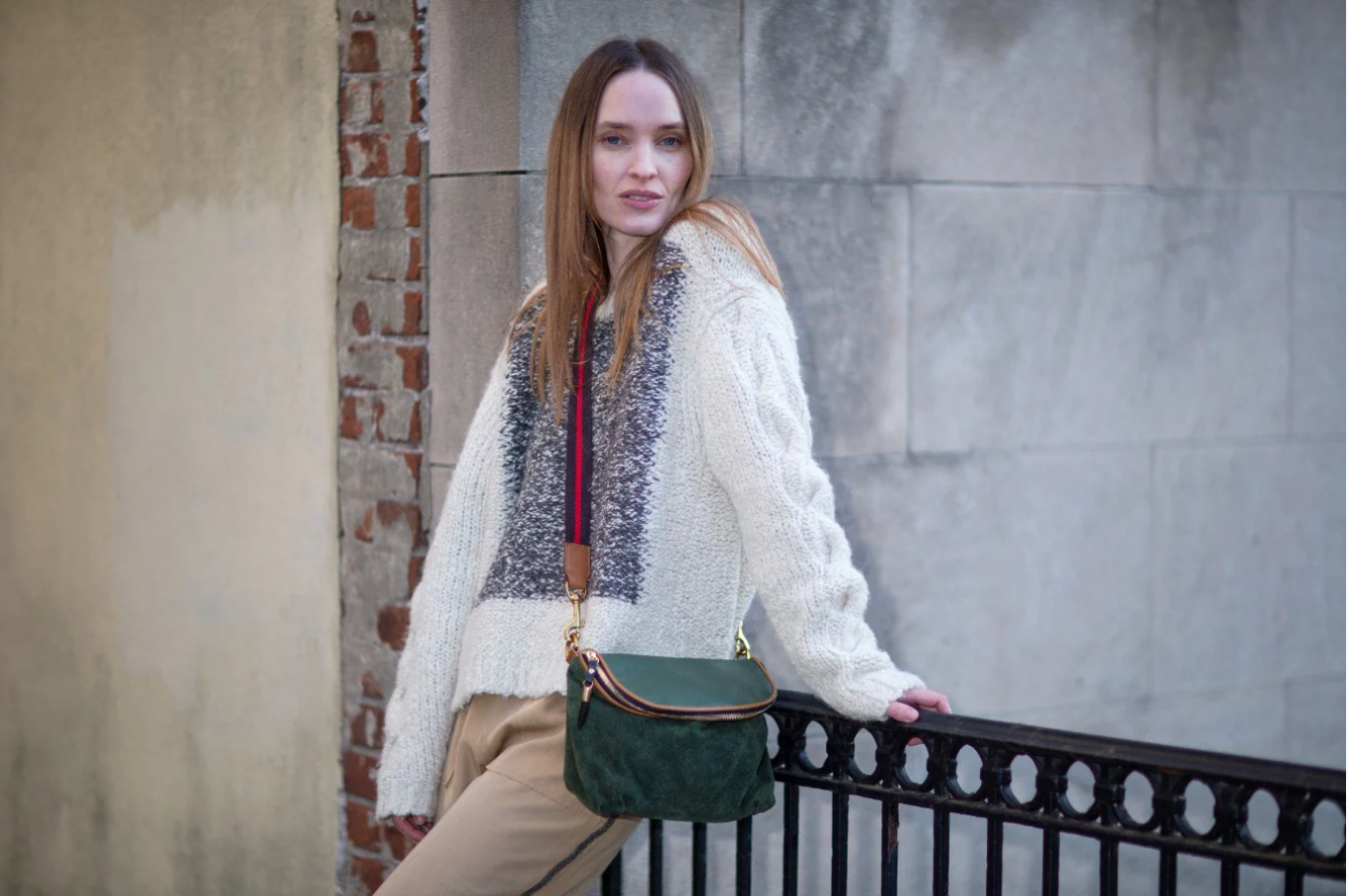 Woman in a white sweater with a patterned vest and green bag standing against a brick wall.