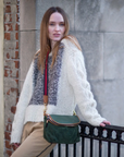 Woman in a white sweater with a patterned vest and green bag standing against a brick wall.
