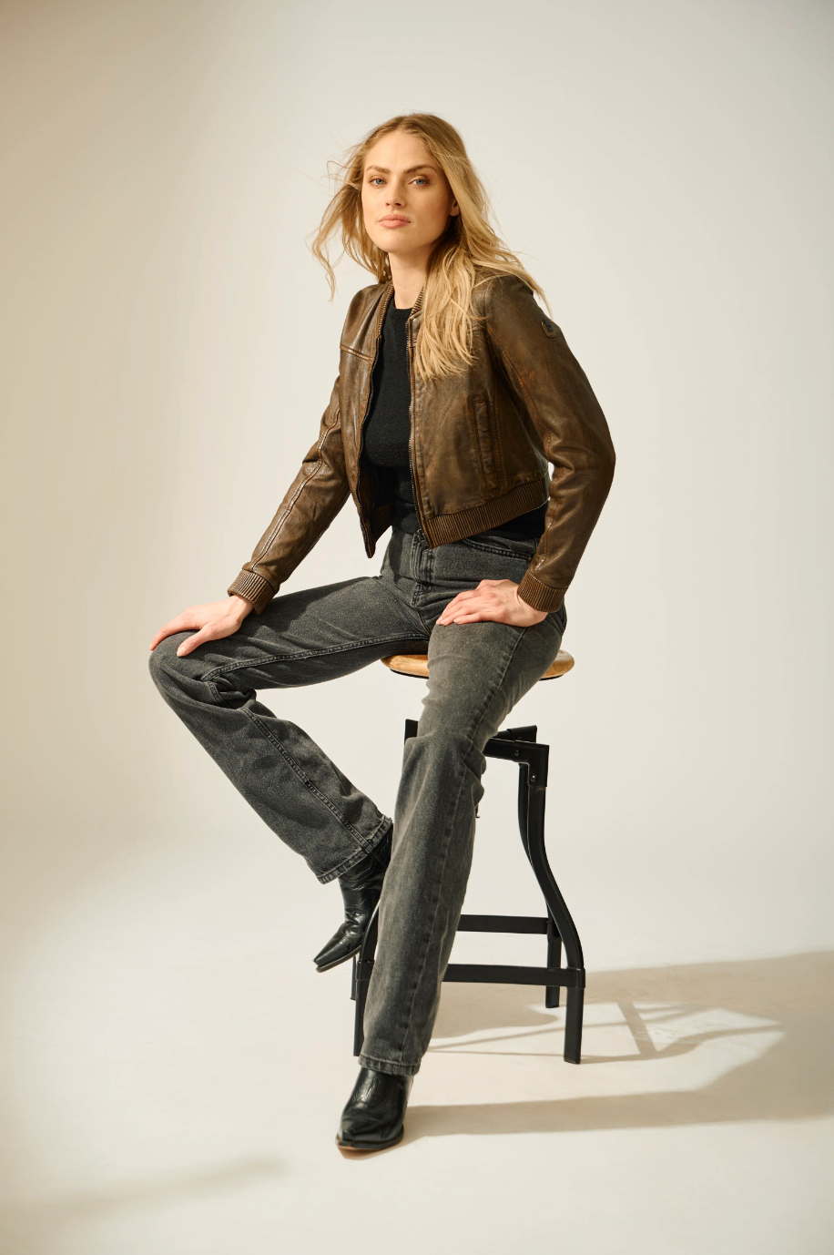 Woman sitting on a stool wearing a brown leather jacket and gray jeans against a plain background