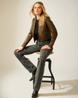 Woman sitting on a stool wearing a brown leather jacket and gray jeans against a plain background