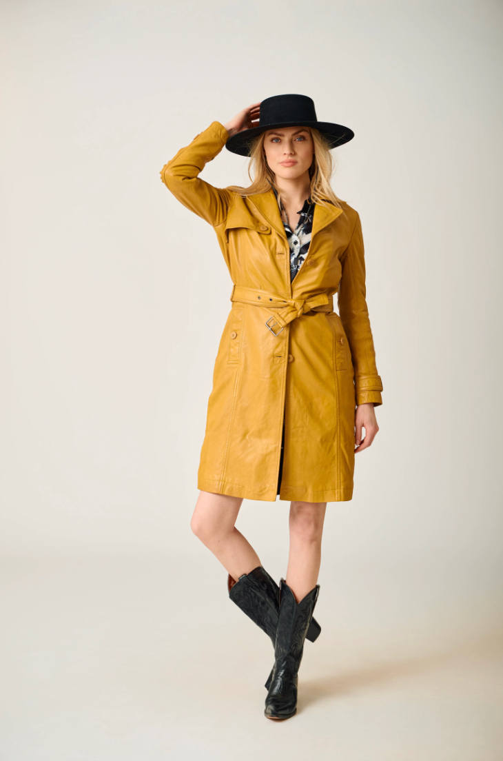 Woman wearing a mustard yellow trench coat, black hat, and black boots on a plain background