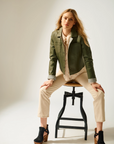 Woman sitting on a stool wearing a green jacket and beige pants against a white background