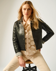 Woman sitting on a stool wearing a black leather jacket over a patterned shirt with beige pants.