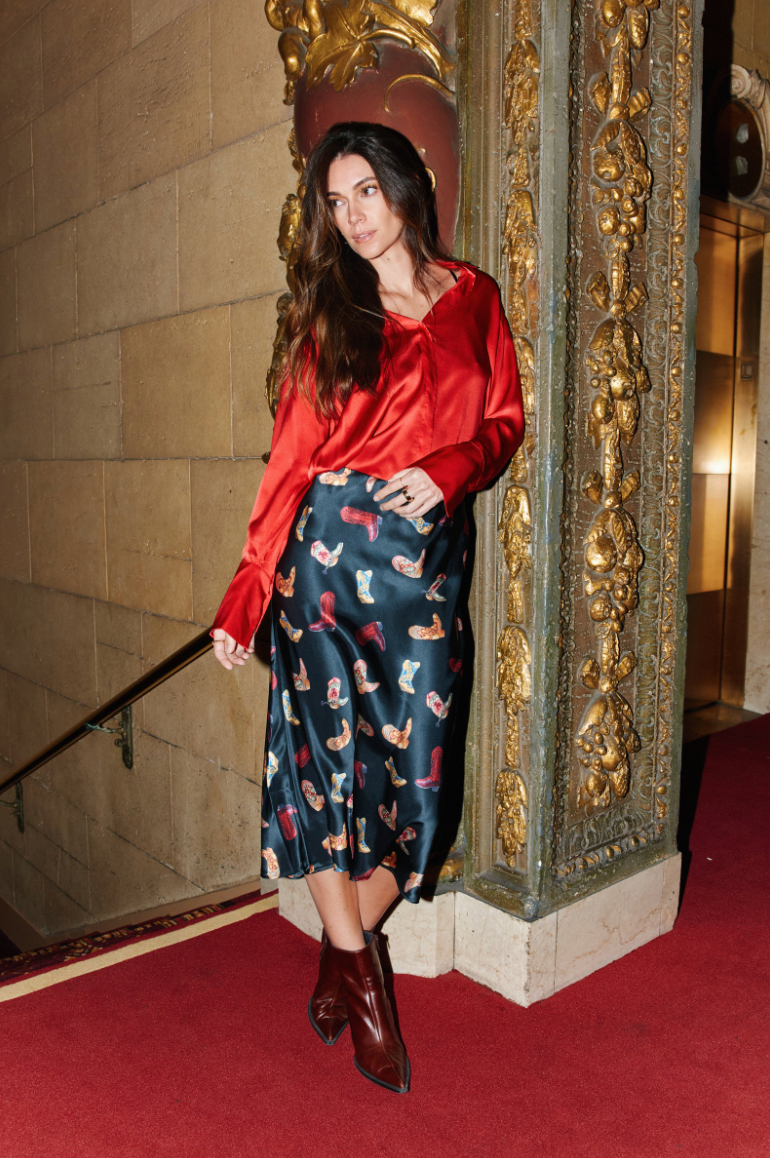 Woman in a red blouse and patterned skirt standing in front of an ornate wall.