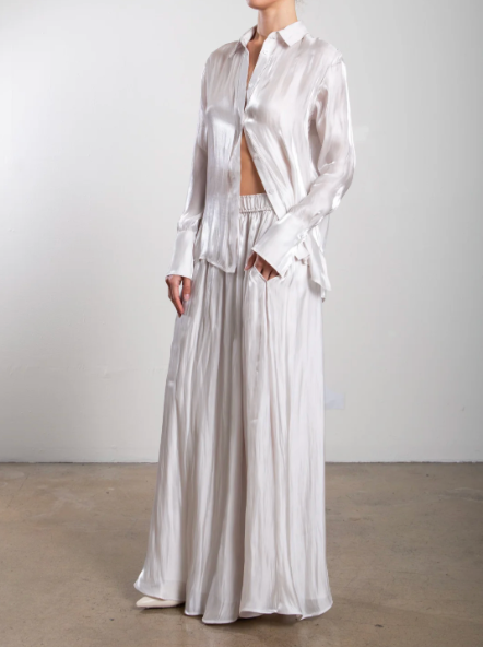 Person wearing a white outfit with a long skirt and open shirt against a plain background