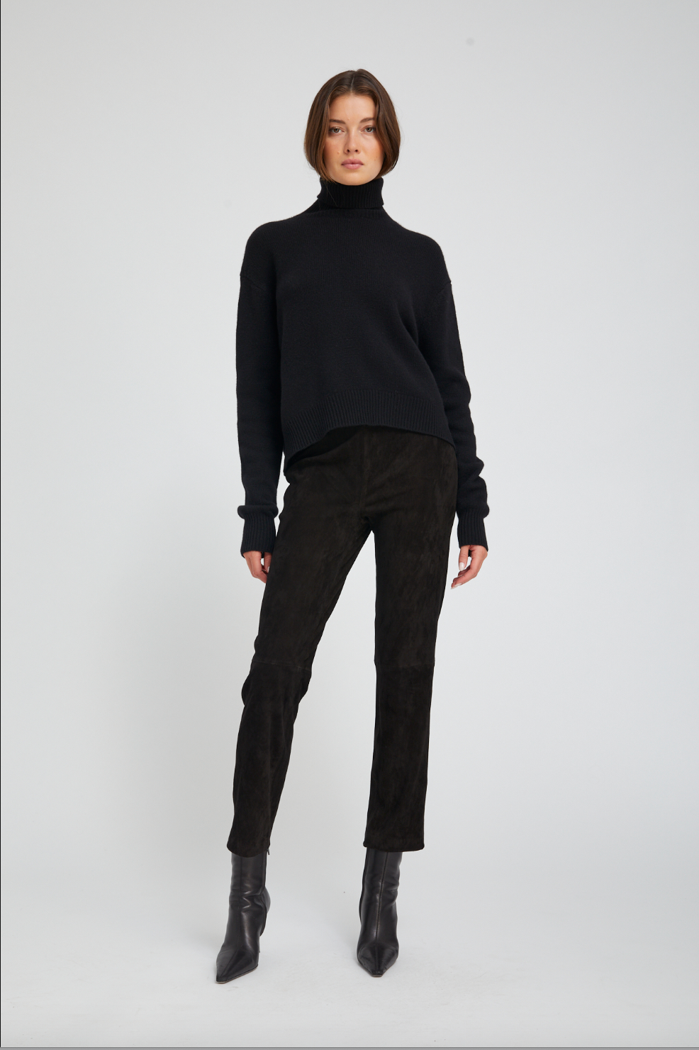 Person wearing a black turtleneck sweater, black pants, and black boots on a white background