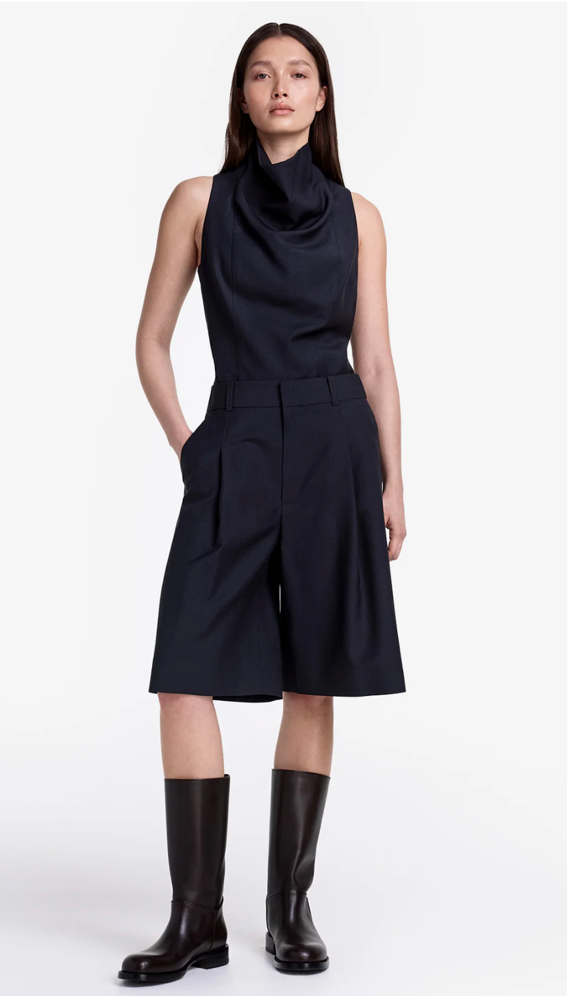Woman wearing a black sleeveless dress with a cowl neck and knee-length skirt, paired with black boots on a white background.